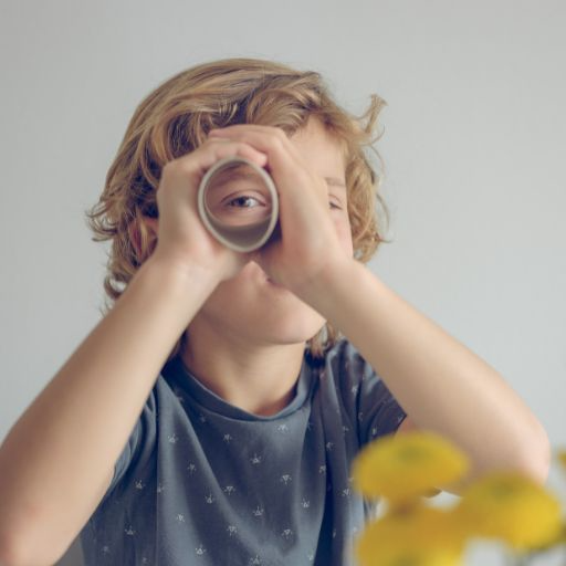 A young, blond haired with curls at their end is wearing a blue T-shirt looking through a paper towel tube using his right eye with a closed left eye. He is holding the tube with both hands. You can clearly see his blue eye looking at you through the opening in the tube.