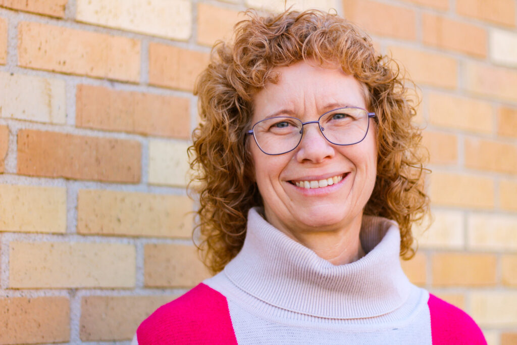 Headshot of Cheri Moore standing in front of a brick wall. She has red, curly hair, blue eyes, and dimples on each side of her smile. She wear glasses and is caucasian. She is wearing a turtleneck cream sweater with red stripe from her shoulders down.
