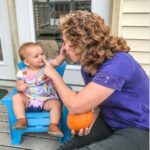 Toddler looking and touching a lady's nose. The lady is touching the toddler's nose and holding a pumpkin. The girl toddler is sitting in a blue chair the perfect height for a toddler.